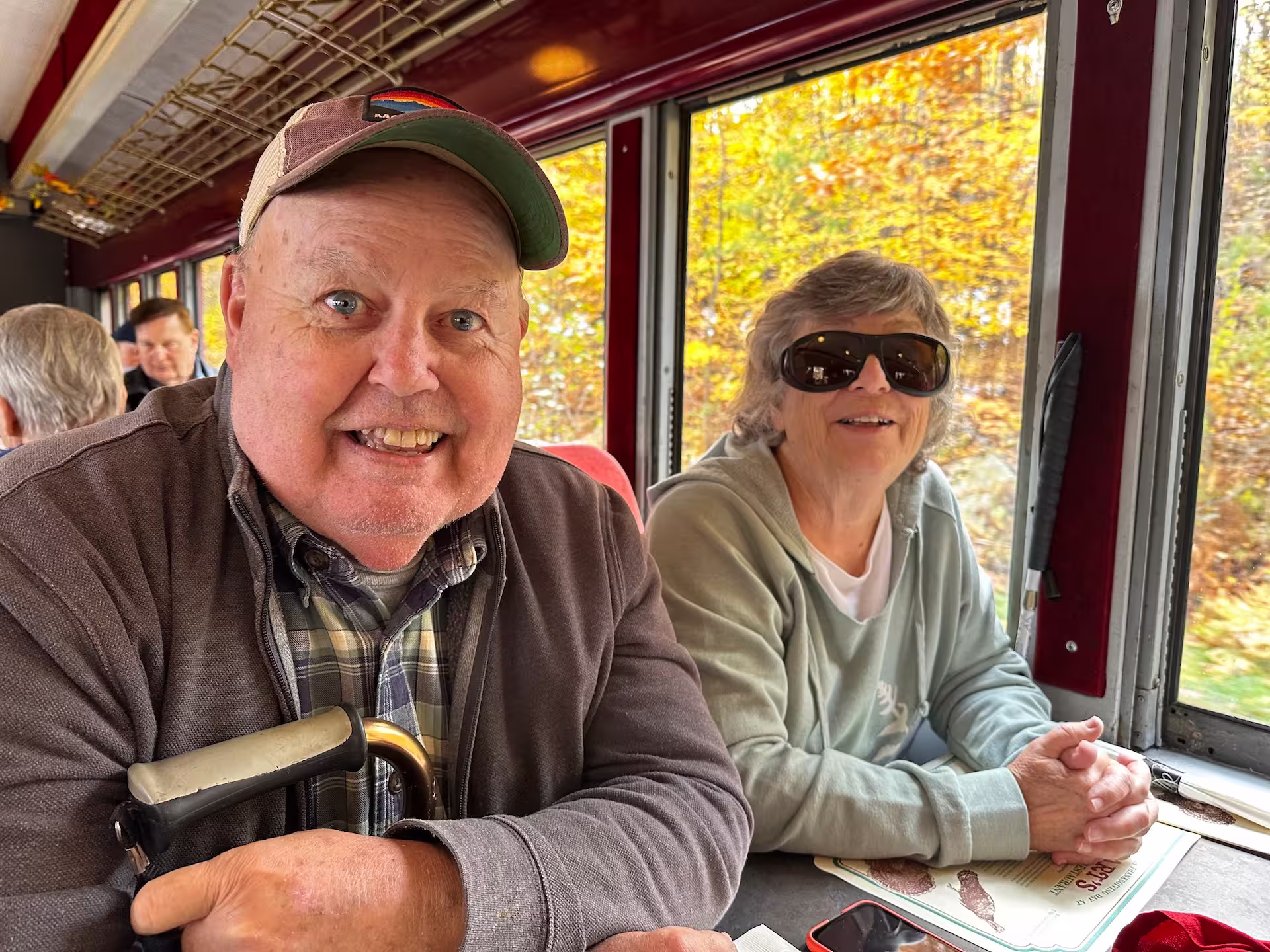 Two people sitting a table with fall foliage visible through the window next to them.