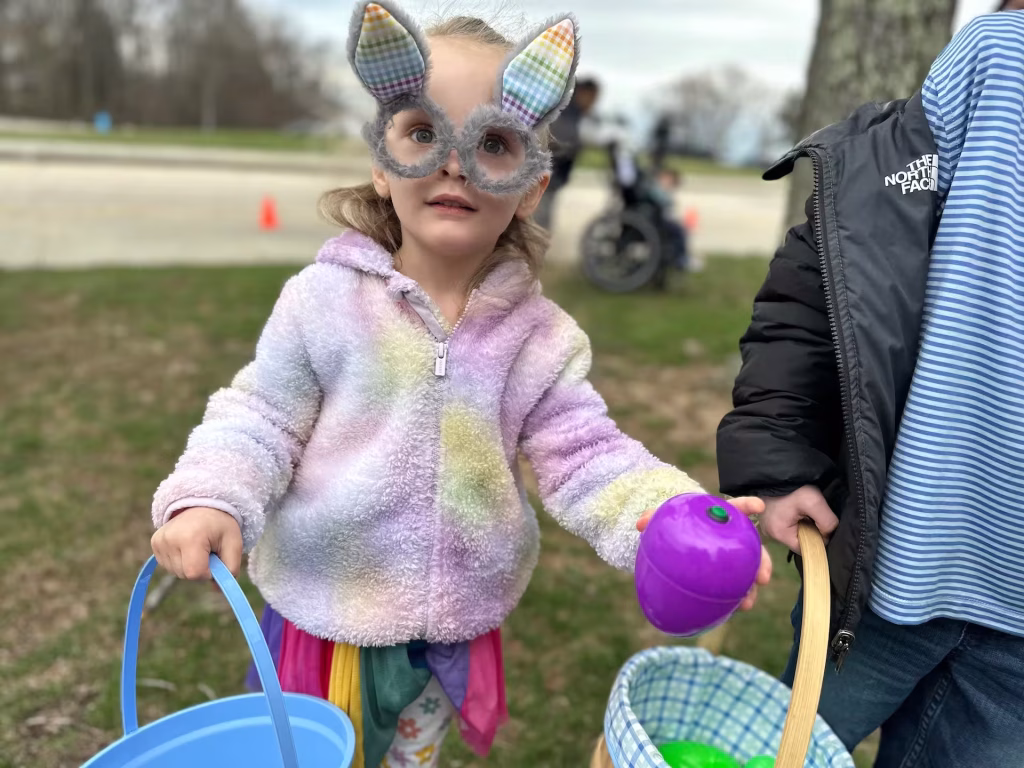 A girl wearing bunny ear glasses holding two baskets and a plastic beeping egg.