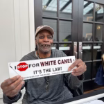 A man holds a sign that says "I stop for white canes. It's the law."