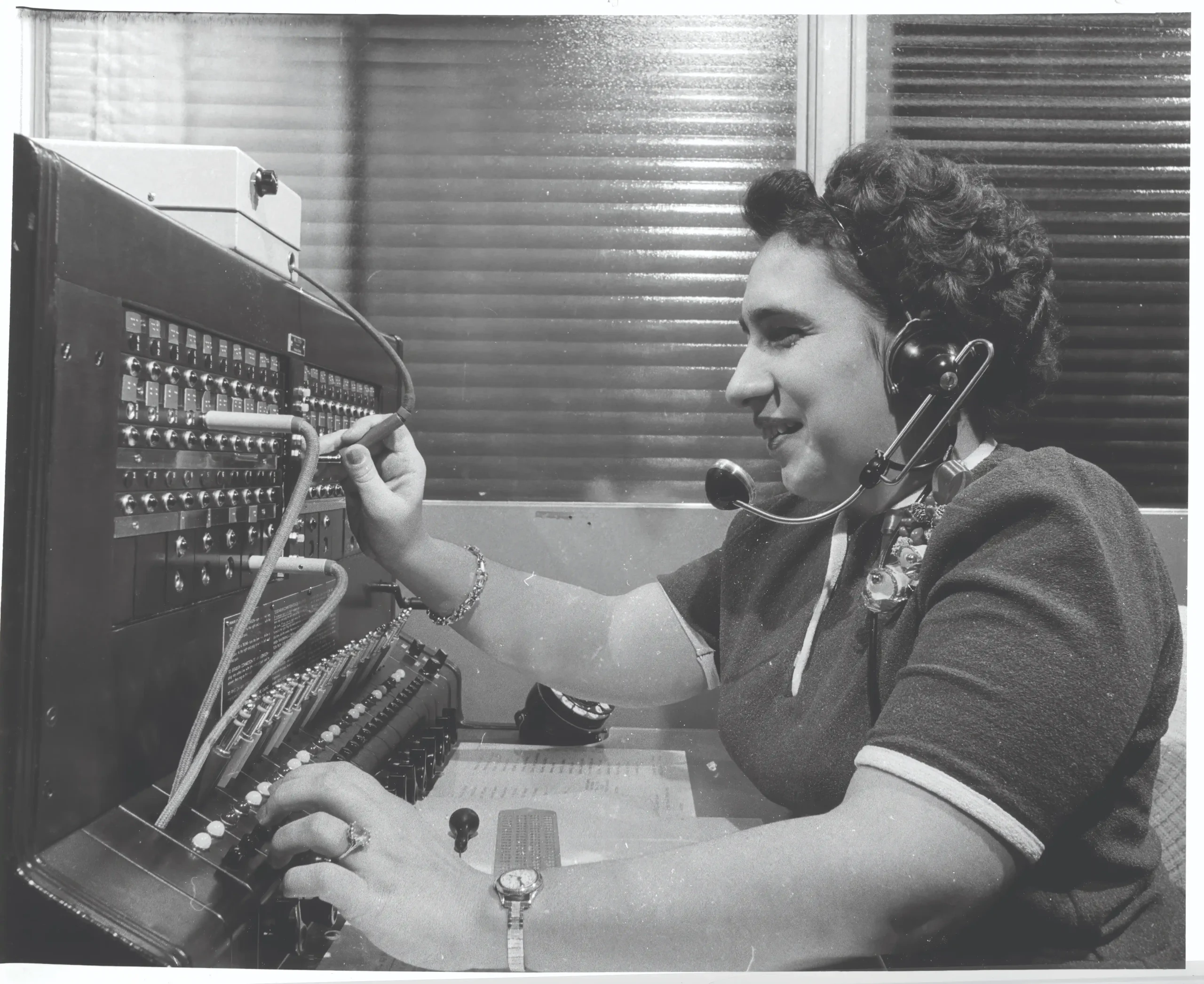 A woman wearing a headset plugs wires into a phone switchboard console.
