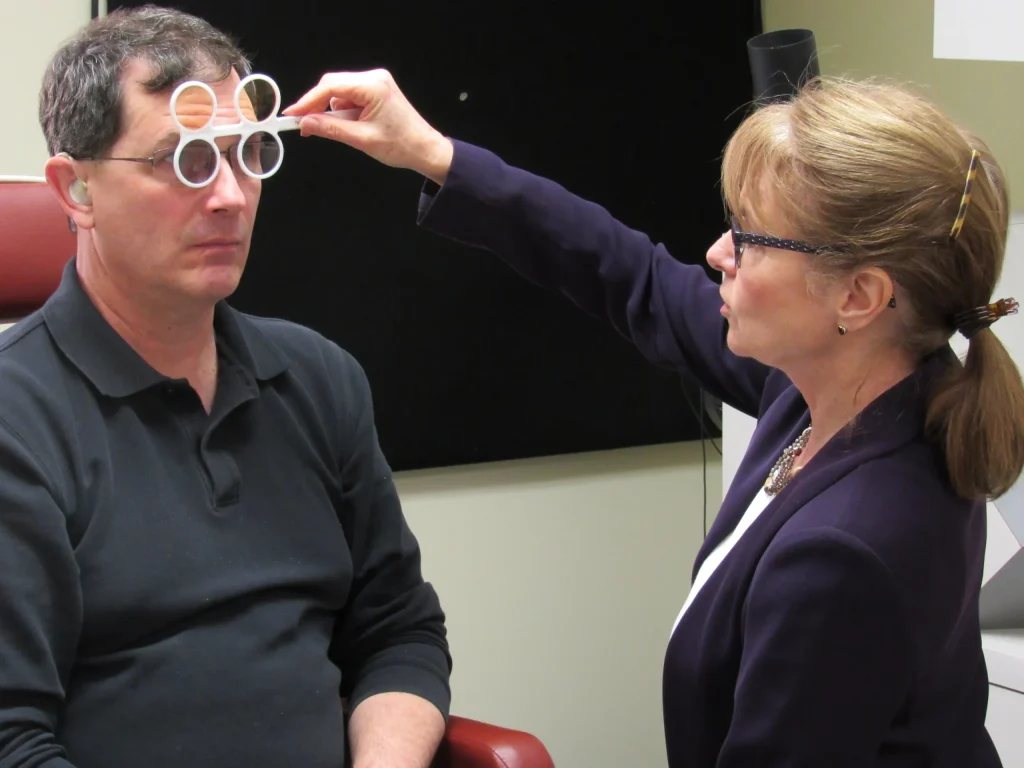 A peron sits in an exam chair while an eye doctor holds tinted glasses up to his eyes.