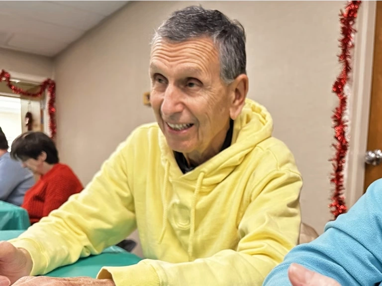 A man wearing a yellow hoodie sweatshirt smiles while sitting a table.
