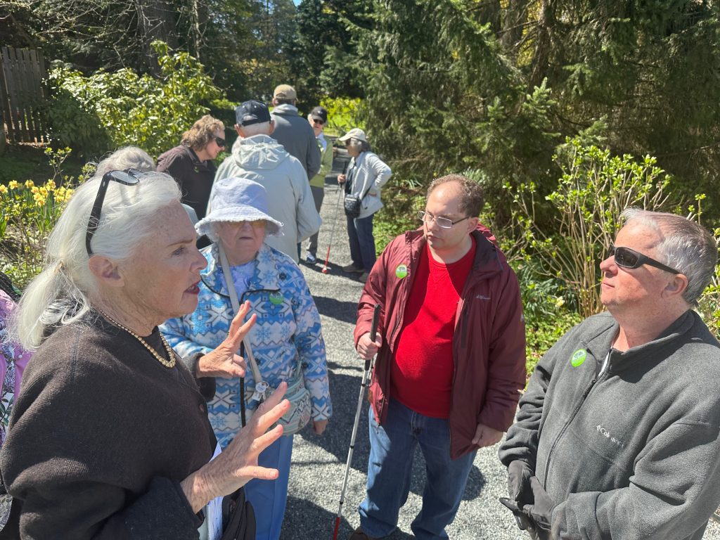 Group of seniors gathered on a sunny garden path, chatting during a guided walk with white canes nearbyy.