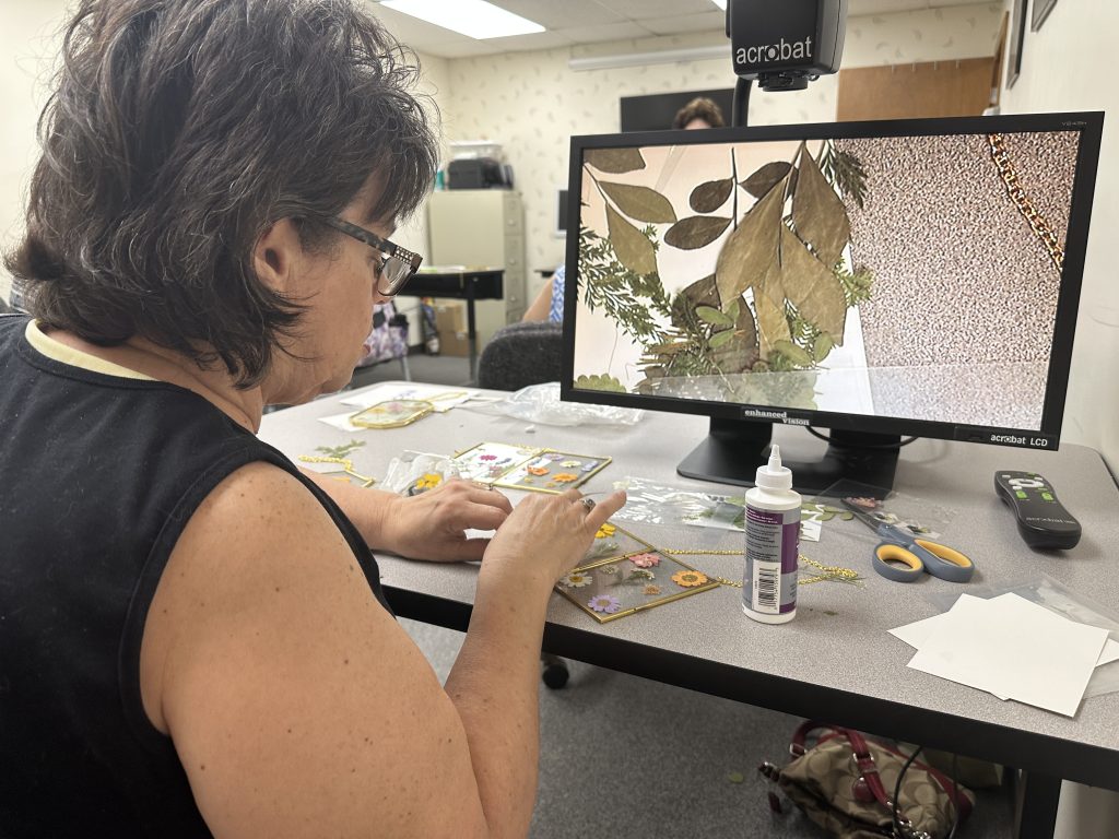 A woman places pressed flowers on a glass frame using tweezers while looking at the image on a video magnifier