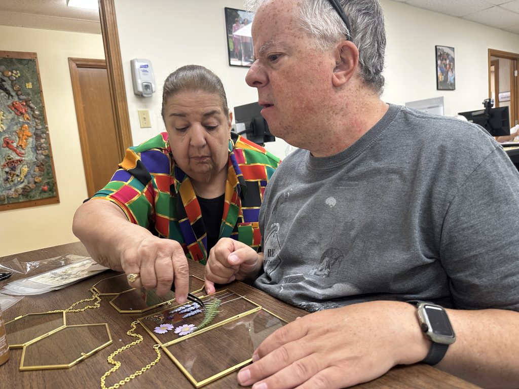 A woman assists a man to place pressed flowers on a glass frame using tweezers